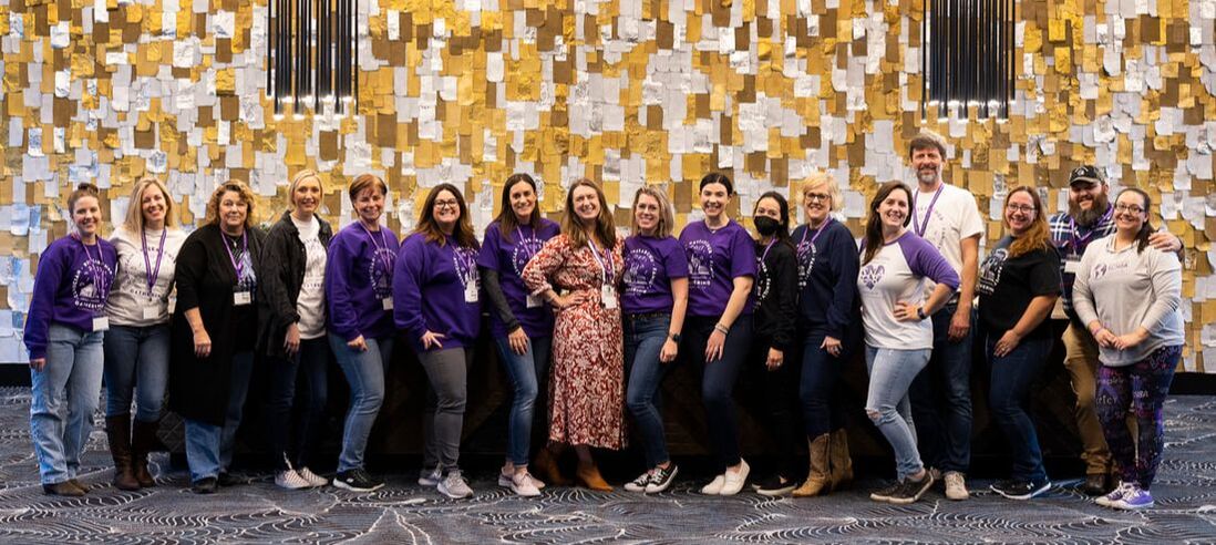 A cheerful group of The Cute Syndrome Foundation volunteers wearing a variety of purple shirts stand in front of a decorative wall embellished with gold and beige accents.
